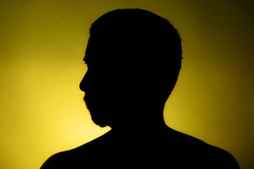 Cleanly defined silhouette of a male person turned to the left against a yellow background with a spotlight and bright area right behind the bust. Studio shot with well defined colour background.
