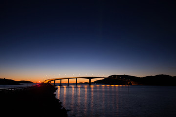 Landscape silhouette and bridge with light on the horizon after sunset. Cloudless, dark blue and symmetrical reflections in the water. Norway nearby Sommaroy.