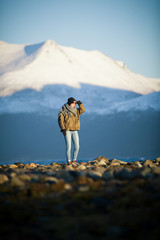 Young woman is standing in the cold landscape of Norway and looking to the sun. Deep camera position with high focal length. In the foreground round stones and in the background snow-capped mountains.