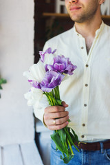 Young male hands holding spring bouquet of purple and white tulips near window in daylight.