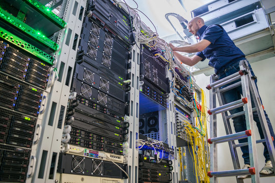 A specialist stands on the ladder in the server room of the data center. Repair of telecommunication equipment. Technician serves computer equipment. Engineer is laying and connecting the wires.
