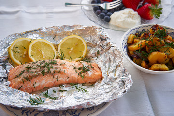 Close up of salmon fillet baked in aluminium foil, slices of lemon and chopped fresh dill served with bowl of roasted potatoes and onions and small plate with fresh blueberries, strawberries and cream