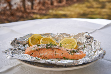 Salmon fillet or steak baked in aluminium baking foil, low fat, fresh and healty dish served as a lunch or dinner in garden restaurant on the whithe plate and slices of organic lemon and chopped dill