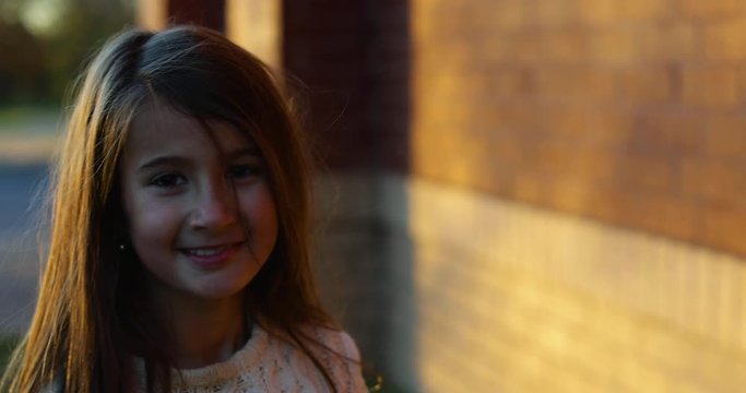 Little girl with straight brown blonde hair in a sweater smiling at the camera while walking backwards in slow motion by a red brick wall during a sunset in winter.