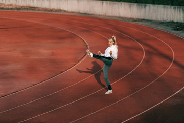 Smiling blonde woman in sportswear kicking in the air while standing on racetrack.