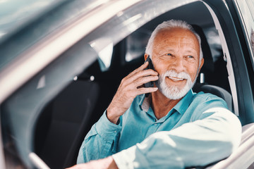 Close up of smiling bearded senior talking on the phone with arm on the opened window while sitting in the car.