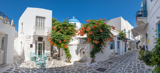 Panorama of the beautiful narrow street in Greece with cozy outdoor cafe and traditional greek church in Parikia town on Paros island, Cyclades - obrazy, fototapety, plakaty