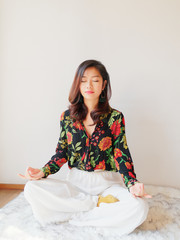 A pretty Chinese young woman practicing yoga at home, sitting on floor with furry cushion against white wall background, exercise, Lotus pose, working out, Feeling peace and wellness concept.