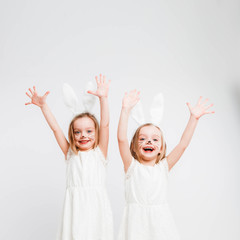 Little blonde twins in white dresses with rabbit ears. Studio photo on gray background. Kids celebrate Easter.