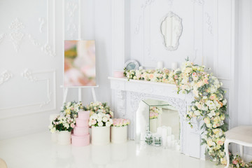 Elegant white fireplace full of flowers. Elegant white room decorated with easel and hat boxes. Wedding decorated area. Vintage decor in light interior