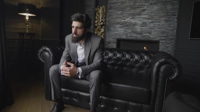 slow motion elegant severe bearded man in grey suit sits on black leather sofa against decorated wall with fireplace
