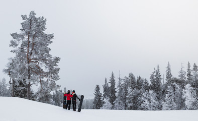 Couple standing toghether on the top of Idre Fjall, Sweden looking down the slope with snow covered trees around them. Getting ready to ride their snowboards down the slope. 
