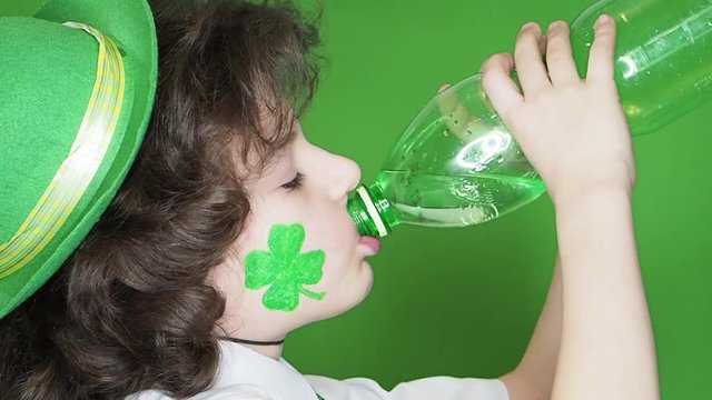 Happy boy with hat and clover image on his cheek drinks water from a bottle isolated on a green background. Profile, Slow Motion.