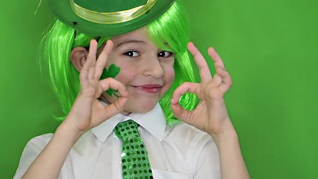 Child Celebrating St. Patrick's Day Showing his Make-up. A small, boy in green wig with leaf of clover and Irish flag on his cheeks showing a gesture okay. slow motion. green background