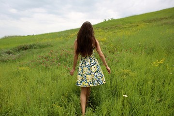 summer walk on a green ravine, a young slim pretty girl with long brown hair in a yellow dress sundress, enjoys life on the field with summer flowers