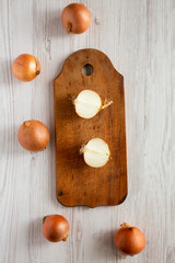 Top view, unpeeled raw yellow onions on rustic wooden board over white wooden surface, top view. Flat lay, overhead, from above.