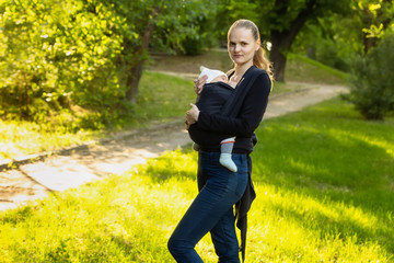 A young mother with her baby in a sling scarf is standing in a park. Newborn carrier. Winding sling for carrying a child