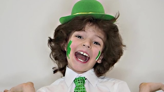 Child Celebrating St. Patrick's Day Laughing and Looking at Camera. A small, curvy boy with drawing in the form of a leaf of clover and Irish flag on his cheeks. slow motion. light background