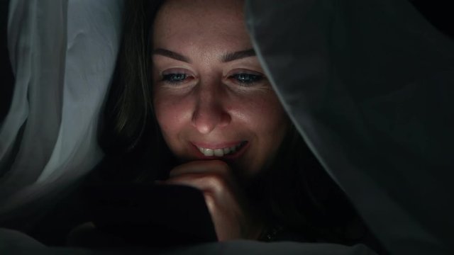 Woman is browsing social media in mobile phone at night under the blanket, Face close-up.