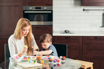 happy blonde mother and four-year old son making homemade Easter eggs, enjoying leisure time spent together at home. Family celebrating Easter. Parent and kid play indoors