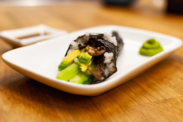 Homemade vegan Temaki-Sushi filled with avocado on a small plate on a wooden table with soy sauce and wasabi