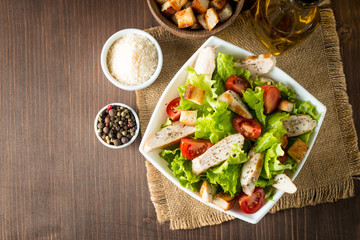 Fresh salad made of tomato, ruccola, chicken breast, eggs, arugula, crackers and spices. Caesar salad in a white, transparent bowl on wooden background