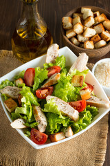 Fresh salad made of tomato, ruccola, chicken breast, eggs, arugula, crackers and spices. Caesar salad in a white, transparent bowl on wooden background
