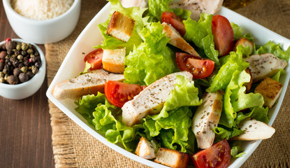 Fresh salad made of tomato, ruccola, chicken breast, eggs, arugula, crackers and spices. Caesar salad in a white, transparent bowl on wooden background