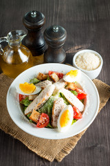 Fresh salad made of tomato, ruccola, chicken breast, eggs, arugula, crackers and spices. Caesar salad in a white, transparent bowl on wooden background