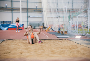 Young blonde smiling women performing a long jump in the sports arena. Sitting in the sand