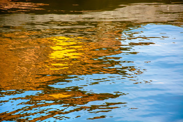 Fototapeta na wymiar Natural watercolors painting concept background. Abstract waving water surface with golden buildings and clear blue sky reflected on the rippling water.