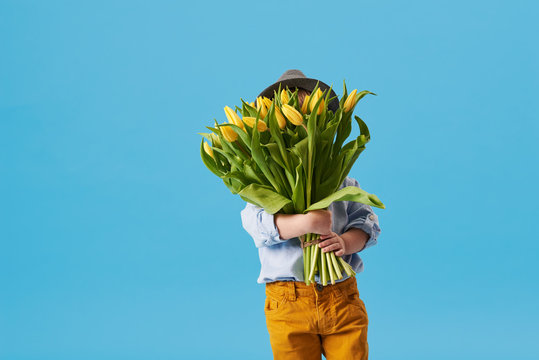 Cute Smiling Child Holding A Beautiful Bouquet Of Yellow Tulips In Front Of His Face Isolated On Blue. Little Toddler Boy Gives A Bouquet To Mom