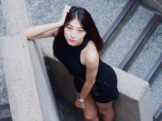 Graceful young woman with long black hair stand casually and lean against the wall, looking at camera emotionally. Outdoor close up photo of cool Chinese girl in trendy summer sexy black dress. 