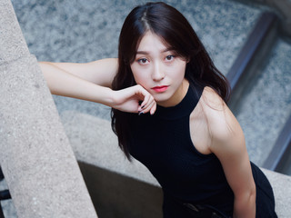 Graceful young woman with long black hair stand casually and lean against the wall, looking at camera emotionally. Outdoor close up photo of cool Chinese girl in trendy summer sexy black dress. 