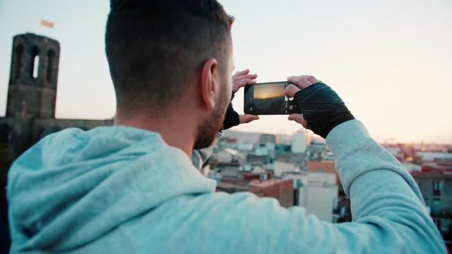 Sport motivated man in good shape taking a smartphone or mobile phone photo or picture of sunset or sunrise on a city rooftop after or before training To capture the moment and apply filter and share