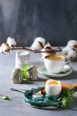 Breakfast with cup of coffee and soft boiled egg, served in green ceramic egg cup with salt, pepper and toasted bread, jug of cream and cotton flowers over grey blue table.