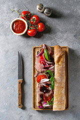 Beef baguette sandwich with tomatoes, basil, red onion served on wooden serving board with ingredients above over grey texture background. Flat lay, space