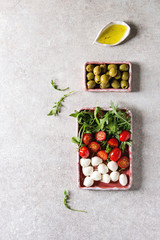 Mozzarella, cherry tomatoes, olives antipasto appetizers served in pink ceramic rectangular plates with olive oil, arugula in rowover grey texture background. Flat lay, space