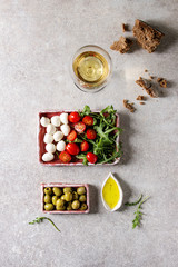 Mozzarella, cherry tomatoes, olives antipasto appetizers served in pink ceramic rectangular plates with olive oil, glass of white wine, arugula, rye bread over grey texture background. Flat lay, space