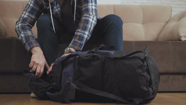 A man sits on the couch, puts things in a backpack, fastens it and leaves the frame.