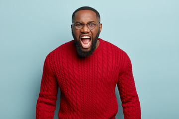 Obraz premium Waist up shot of outraged black man yells angrily with widely opened mouth, expresses negative emotions, dissatisfied of being cheated, wears red knitted jumper, models over blue studio wall