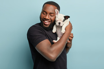 Sweet moments with domestic animals. Portrait of satisfied dark skinned man holds cute small french dog puppy closely to face, enjoys good time with loyal pet, wears casual black t shirt, pose indoor - Powered by Adobe