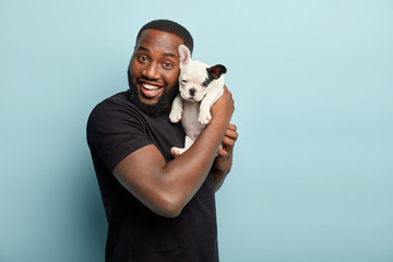 Happy satisfied smiling dark skinned man carries small pet of french bulldog breed, spend leisure time together, expresses positive emotions during photoshoot with dog, isolated over blue background - Powered by Adobe