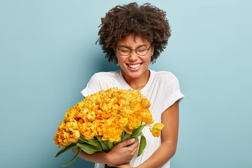 Image of pleased emotional Afro American lady carries yellow tulips closely to herself, giggles at...
