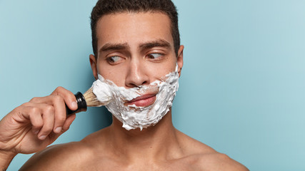 Puzzled naked man has foam on face, keep gaze aside, shaves bristle, wants to have smooth skin, stands with bare shoulders against blue background, blank space on right side. Skin care concept