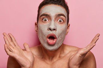 Men beauty and spa concept. Shocked young man stunned to hear big price on cosmetic procedure in salon, spreads hands, has no money to pay cosmetologist, healthy soft skin, nourishing clay mask