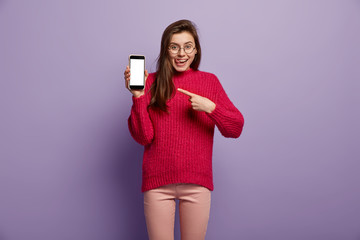 Indoor shot of cheerful lady promots cool gadgets, points with fore fingers at mock up screen of mobile phone, being in high spirit, shows how to use modern electronic device, demonstrates functions