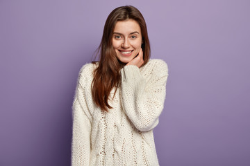 Portrait of tender satisfied model bites lower lip, has toothy smile, keeps hand near cheek, expresses positiveness, hears pleasant news, dressed in white jumper, isolated over purple studio wall