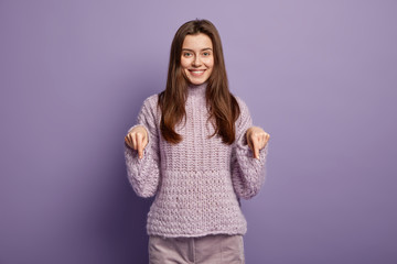 Gorgeous smiling young woman points down on floor, wears knitted sweater and corduroys trousers,...