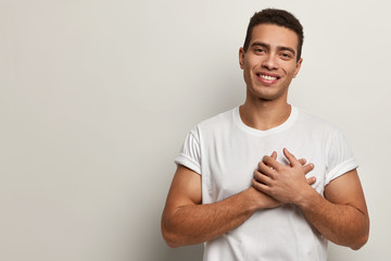 Indoor shot of handsome man keeps hands on chest, being cordial and friendly, expresses gratitude...